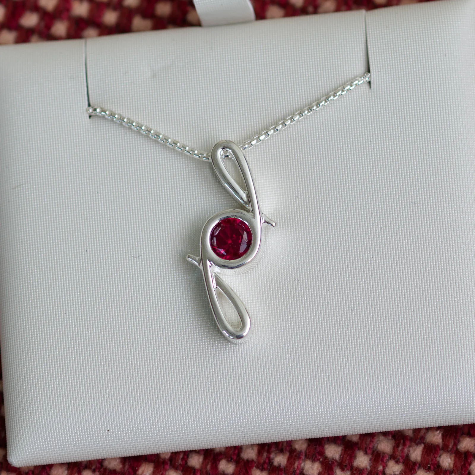 This pendant is made from tarnish resistant silver and features a simulated ruby birth stone. It hangs from a 1mm rounded box chain. The design is a signature motif that symbolizes our message that every life is a precious life.
